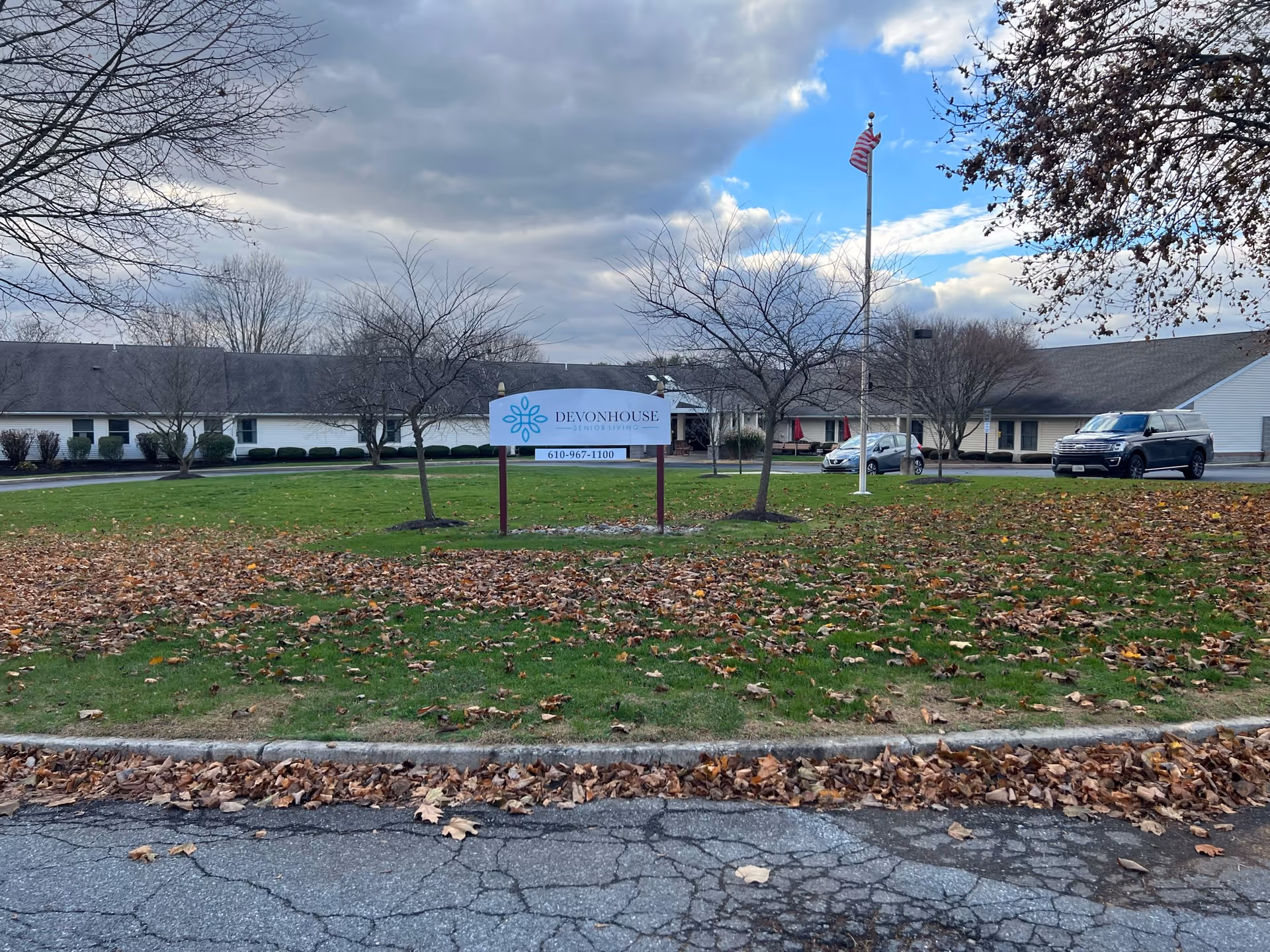 Exterior view of DevonHouse Senior Living Allentown facility with a large sign displaying the facility name and phone number. The building is single-story with a gray roof, surrounded by leafless trees and a lawn covered with fallen leaves. An American flag is flying on a flagpole near the sign, and two cars are parked near the entrance.