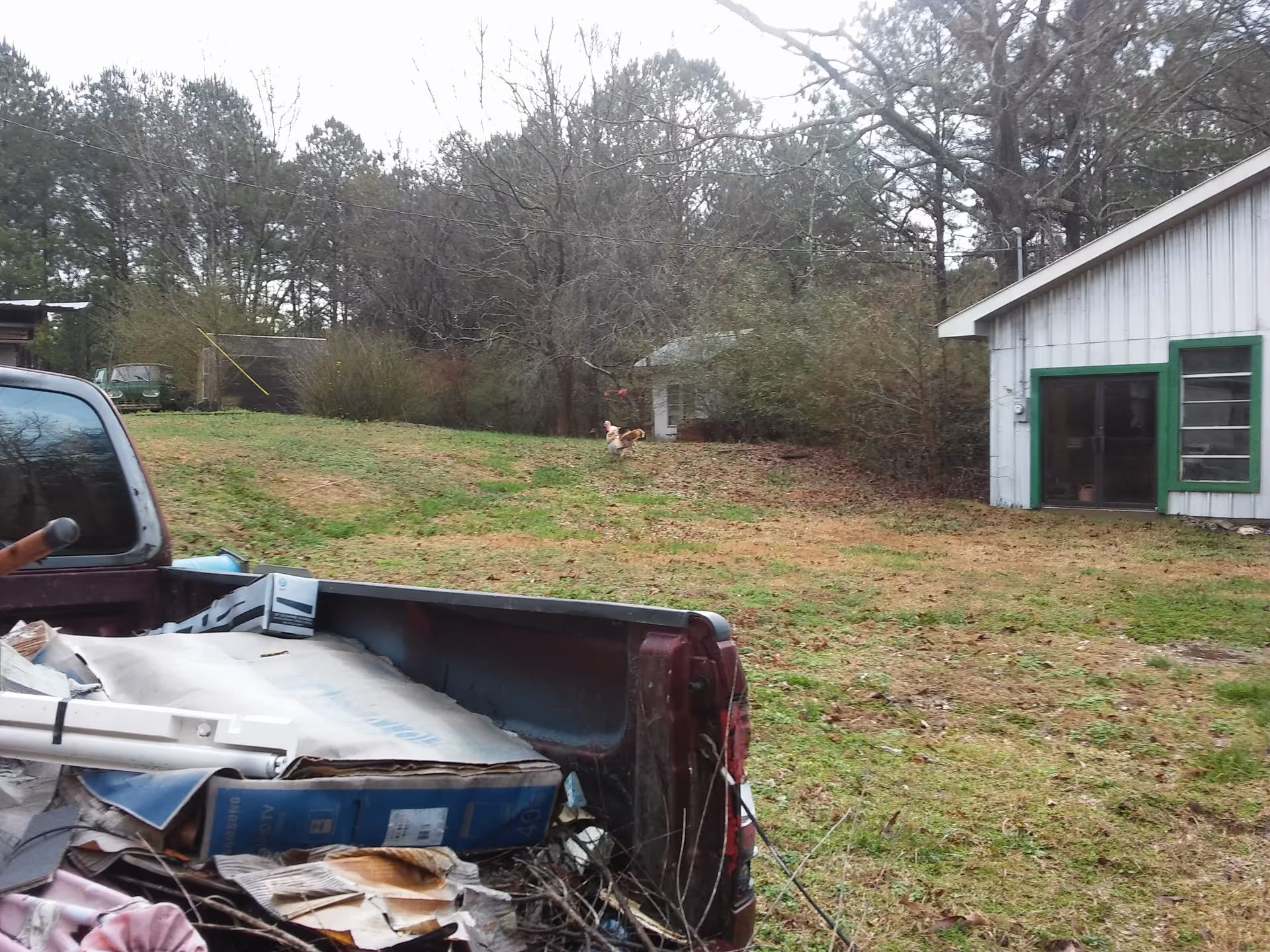 Rugged rural yard with a white building, green trim, and a truck bed filled with debris.