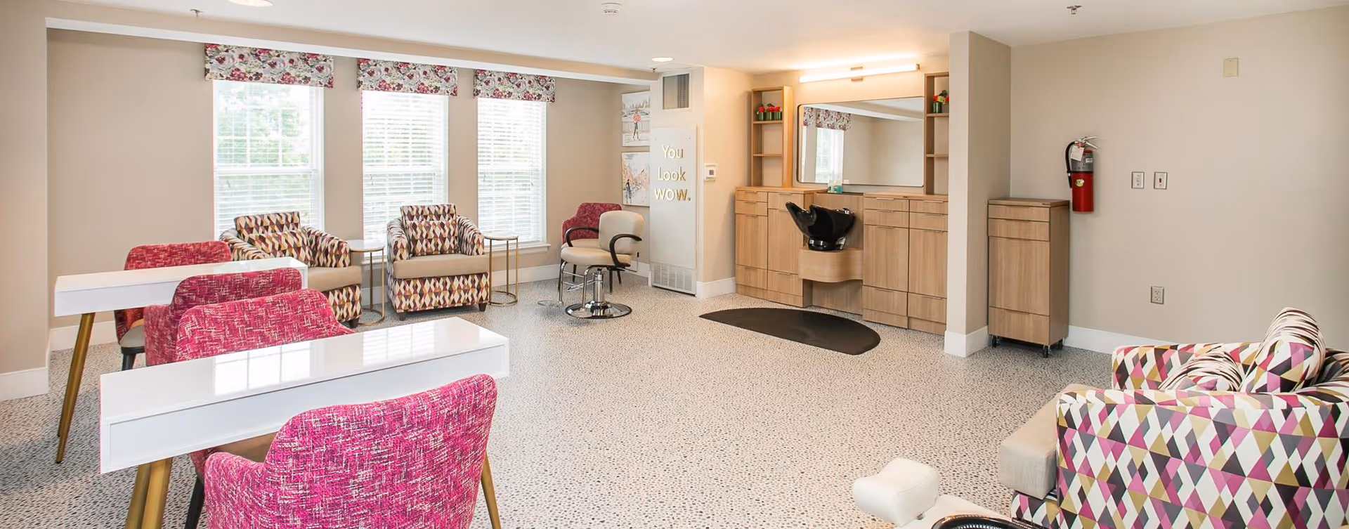 A bright and clean salon area in a senior living facility with multiple colorful chairs, white manicure tables, a hair washing station with a large mirror, and windows with floral valances letting in natural light.