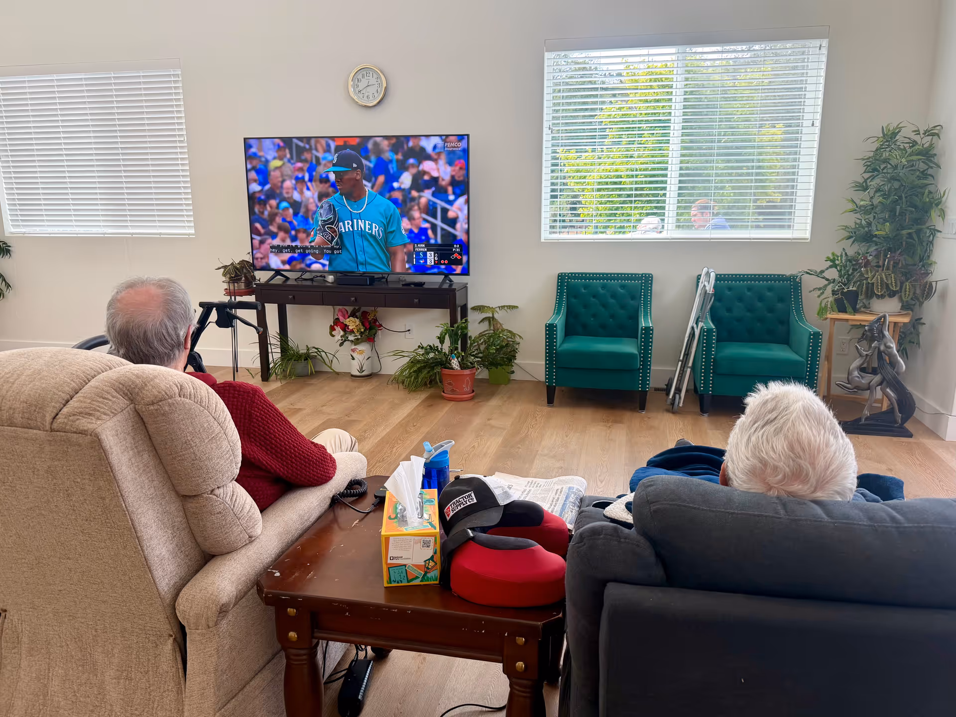 Senior residents lounge in a furnished living room watching TV in a bright common area.
