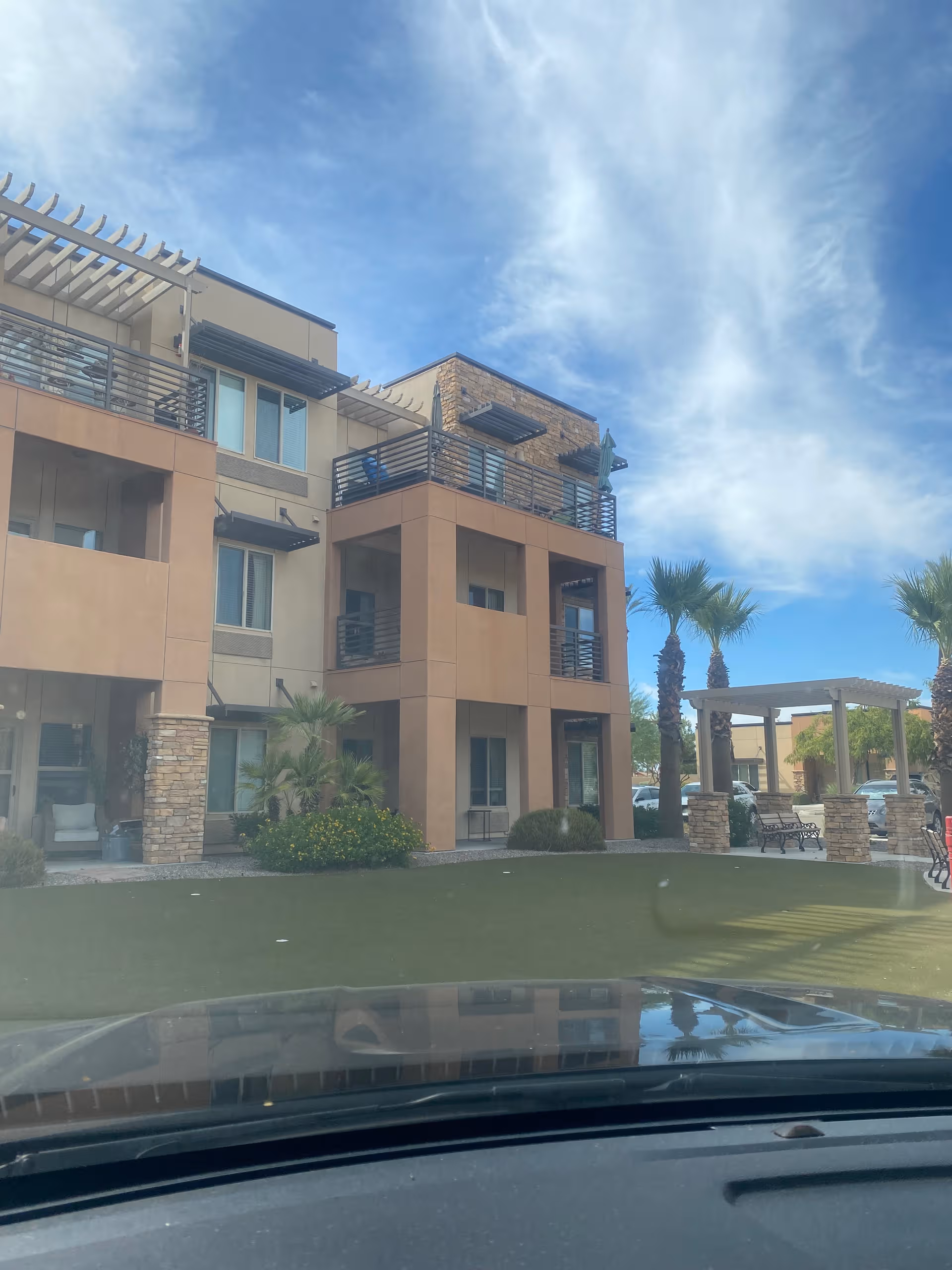 View of a modern, multi-story senior living building with balconies, palm trees, and a seating area.