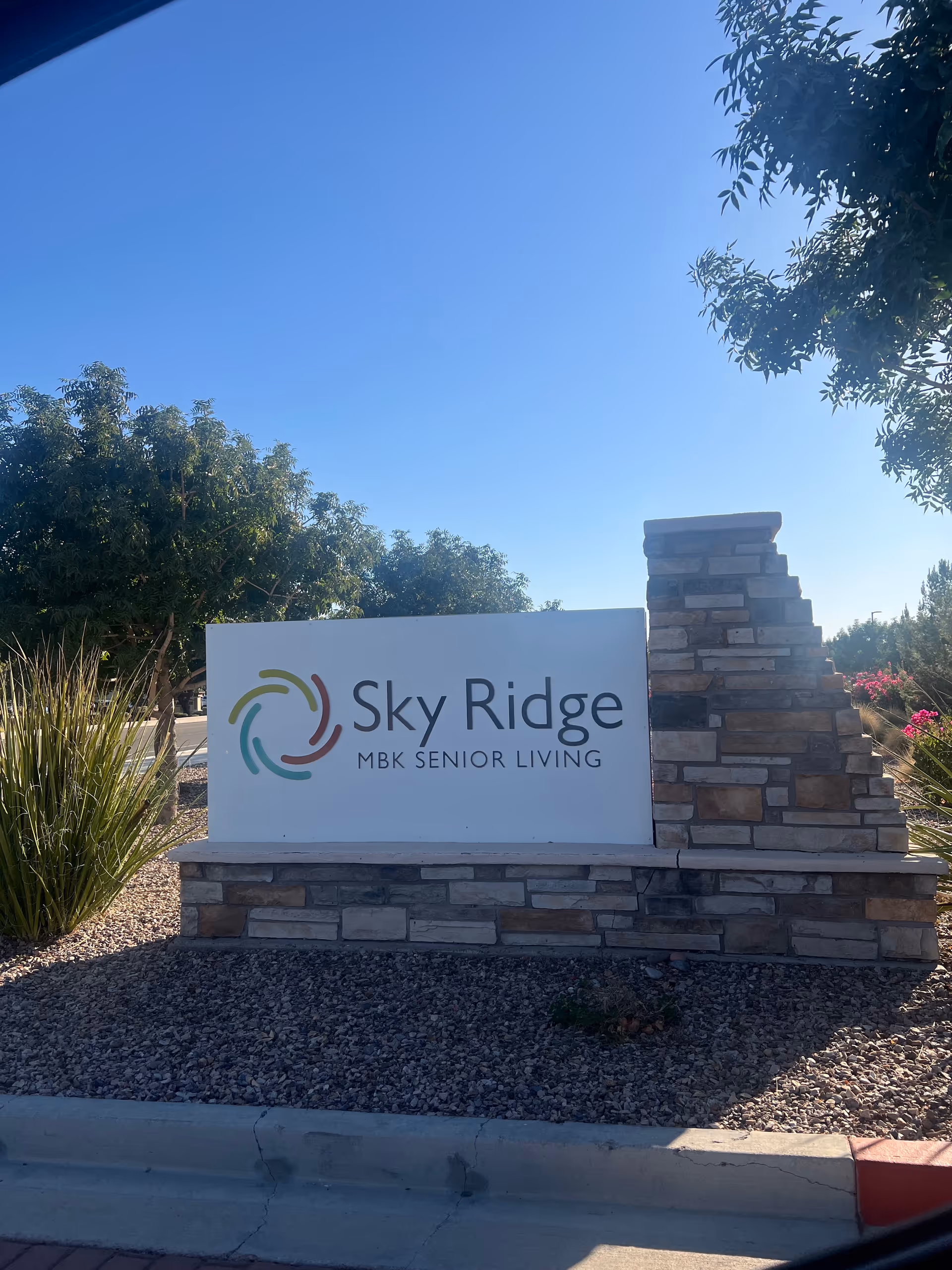 Sky Ridge Senior Living entrance sign with stone pillar and landscaping under a clear blue sky.
