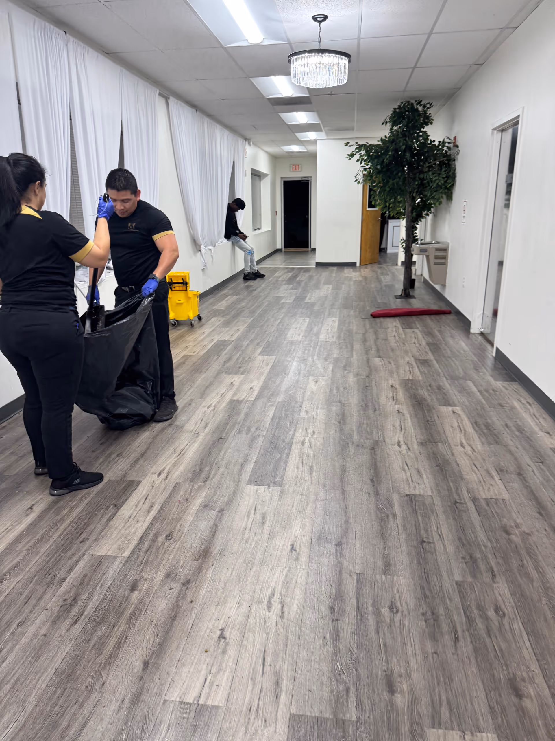 Long interior hallway of a senior living facility with staff cleaning, curtains, and a potted plant.
