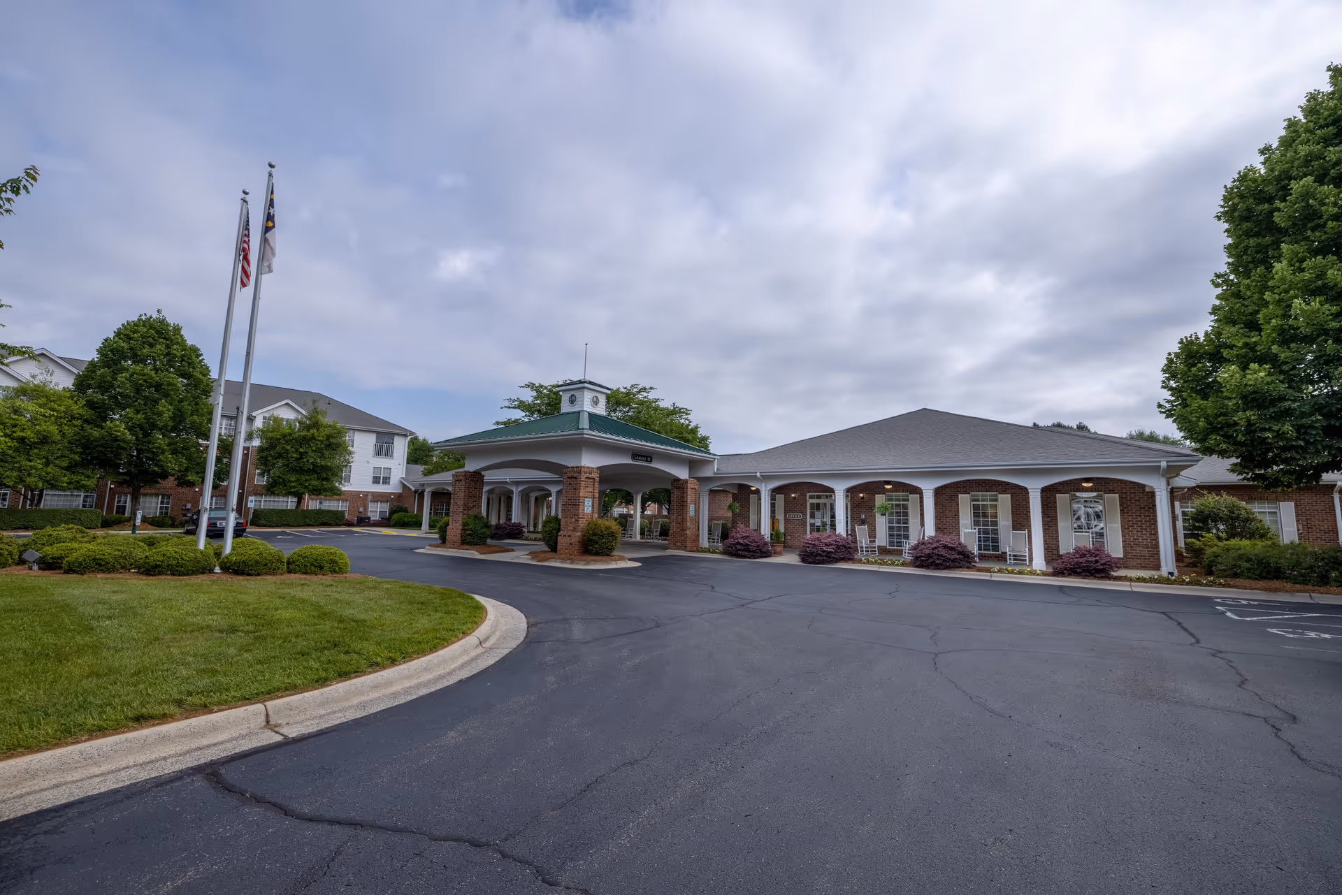 Exterior view of RidgeCrest senior living facility showing a driveway leading to a covered entrance with brick columns and a green roof. The building is surrounded by well-maintained landscaping with trees and bushes, and two flagpoles with flags are visible on the left side.