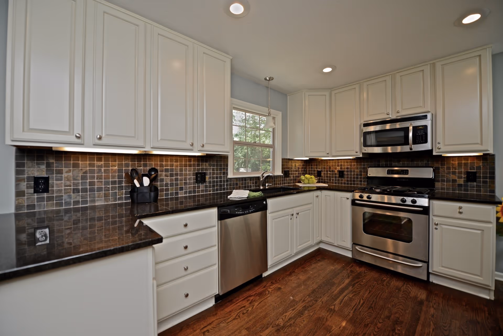 Bright kitchen with white cabinets, stainless steel appliances, dark tile backsplash and hardwood floors.