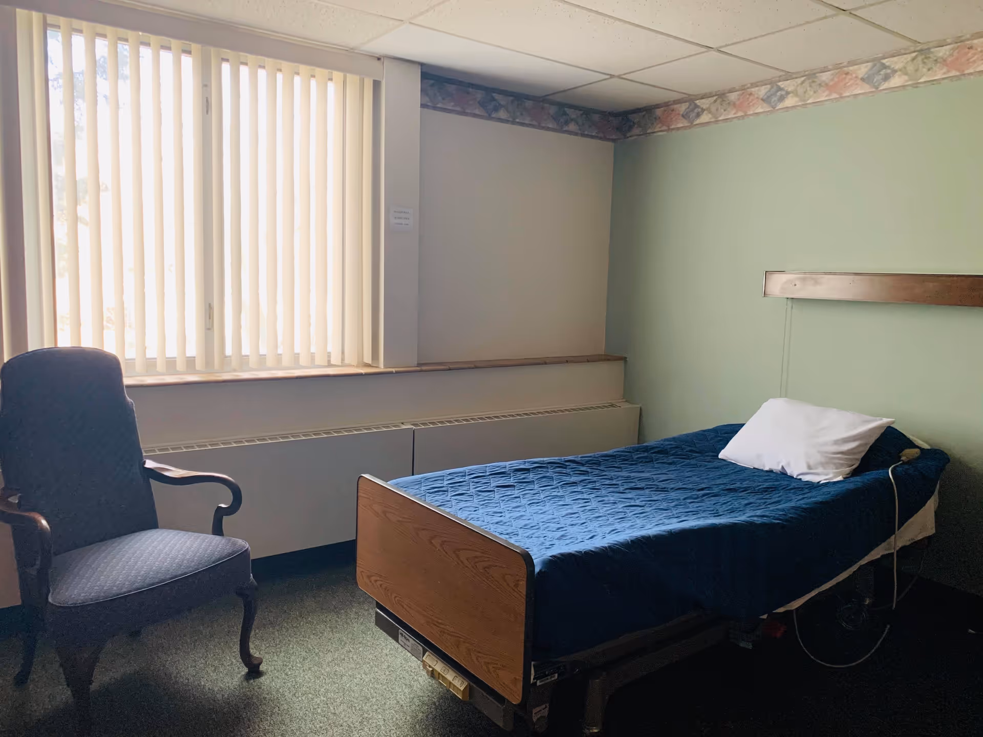 A small room with a single adjustable bed covered in a blue quilt, a pillow, an upholstered chair, and a window with vertical blinds.