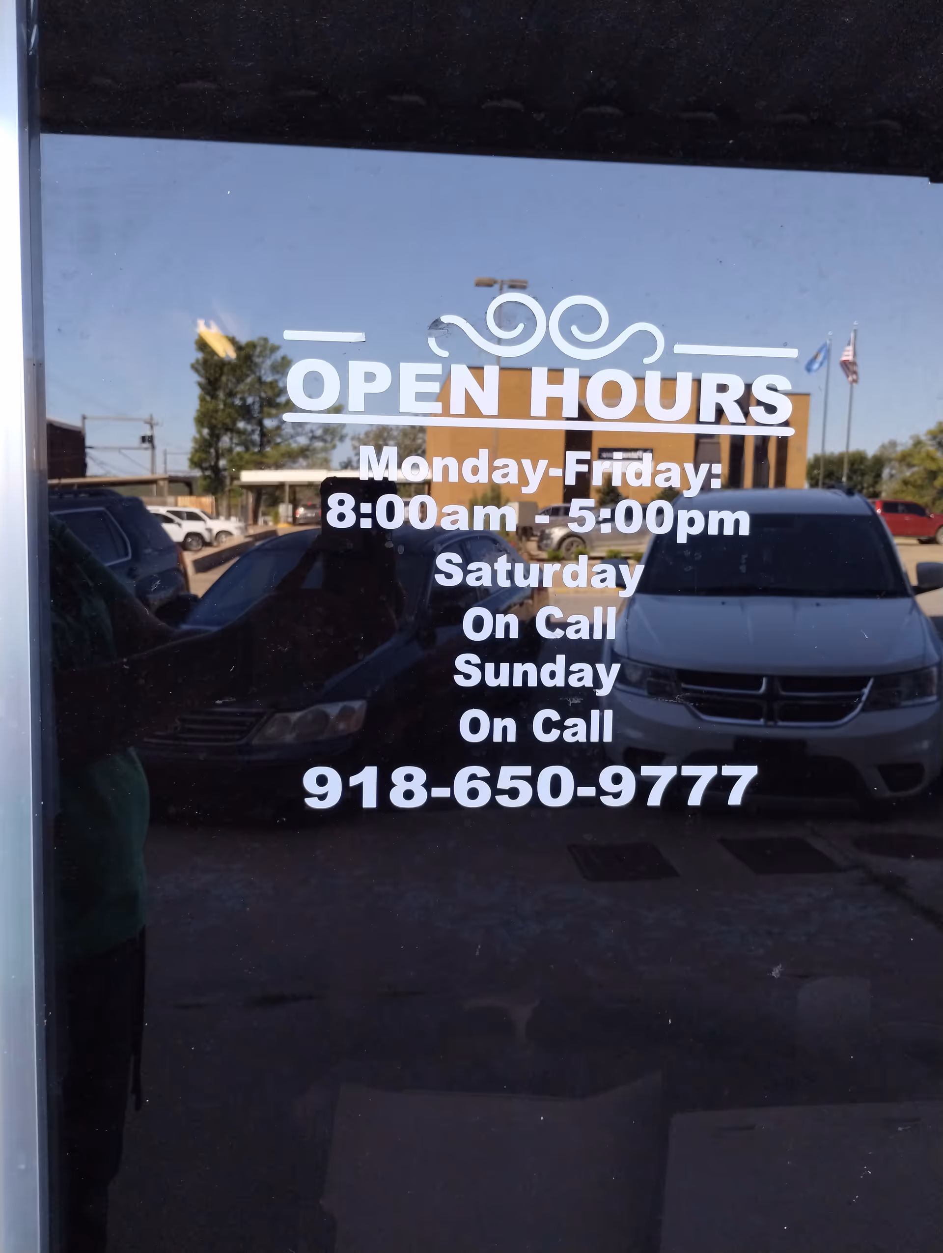 Glass door displays open hours sign with phone number, reflecting parked cars.