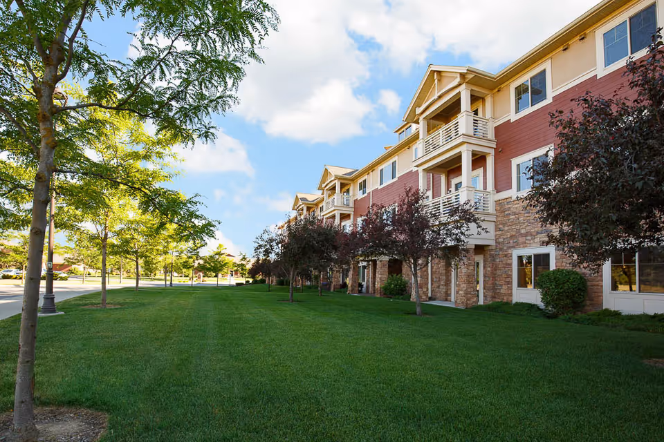 Exterior view of a senior living facility building with a well-maintained green lawn and trees along the side. The building has multiple floors with balconies and a combination of stone and red siding on the facade under a partly cloudy blue sky.