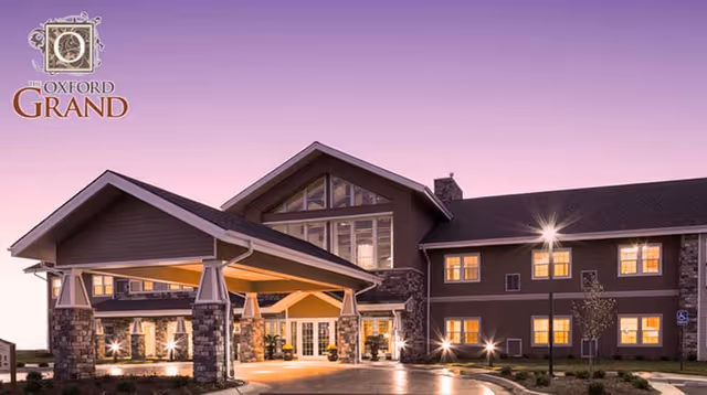 Exterior front of The Oxford Grand assisted living building at dusk with an illuminated porte-cochère entrance and a logo in the top-left corner.