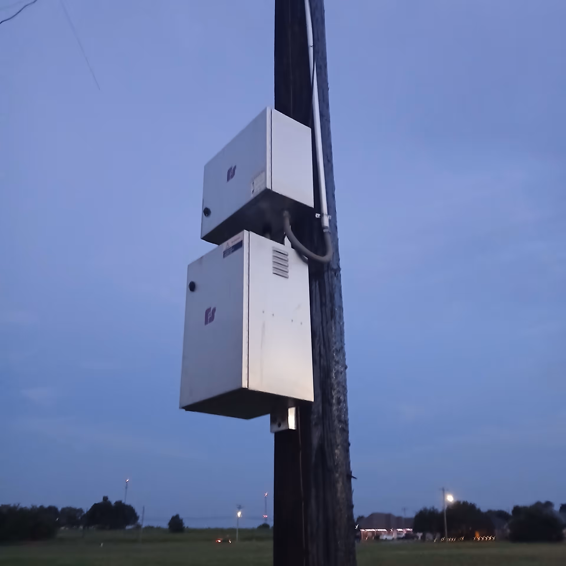 Two white utility boxes mounted on a pole against a blue evening sky and open field.