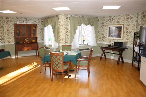 Sunny communal dining room with a round table covered in a green tablecloth, wooden chairs, floral wallpaper, and windows with green valances.