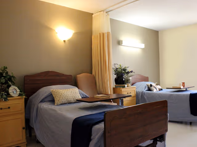 A senior living facility bedroom with two single hospital-style beds, each with blue bedding and a pillow. Between the beds is a small wooden nightstand with a vase of flowers and some books. There is a privacy curtain partially drawn between the beds. The room has soft lighting from wall-mounted fixtures and a window letting in natural light.