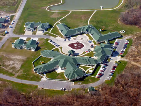 Aerial view of a senior living facility named River Hills Village, showing multiple connected buildings with green roofs arranged around a circular driveway and parking lots, surrounded by grassy areas and trees.