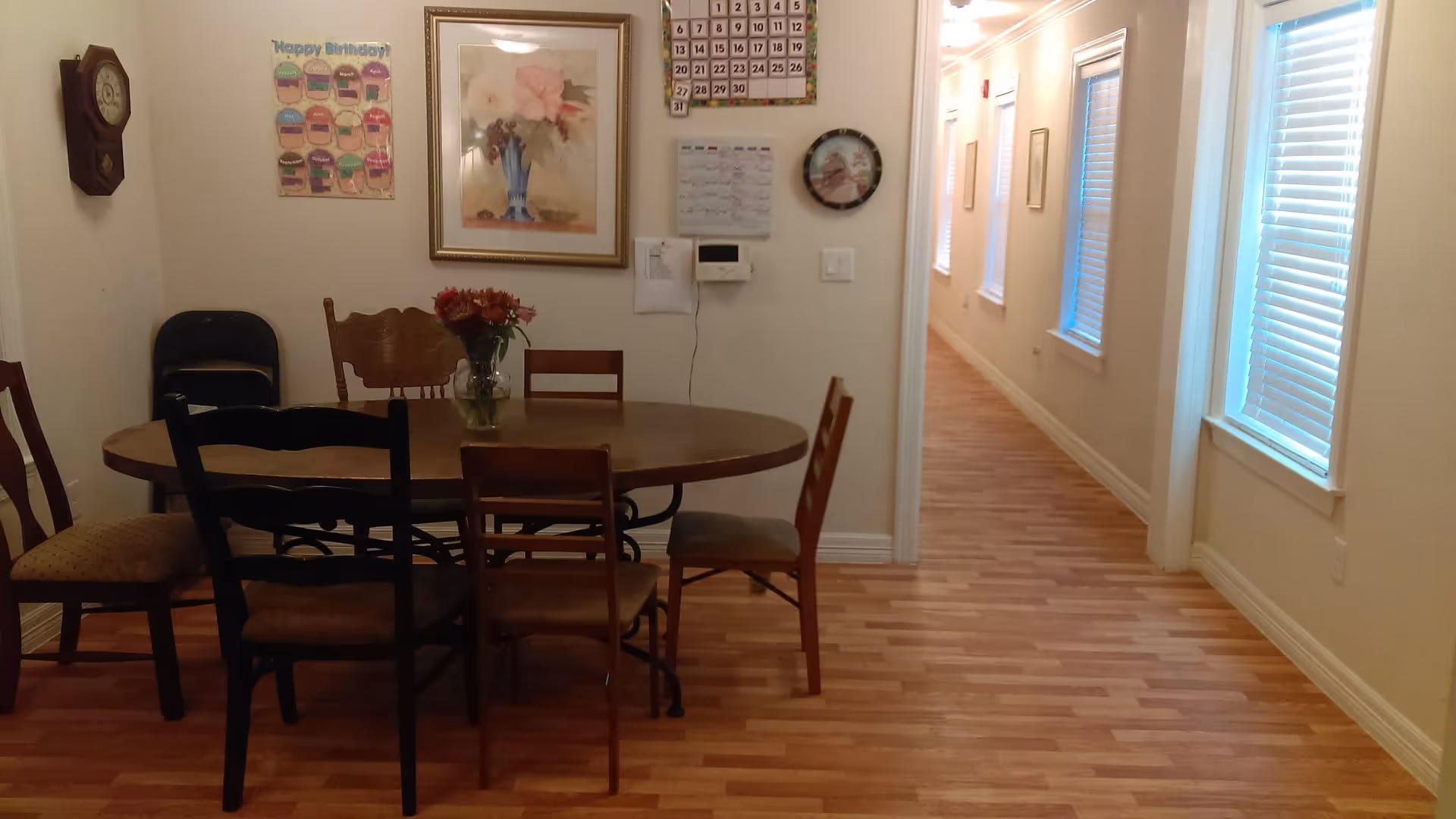 Interior view of a dining area with a wooden oval table surrounded by six chairs of different styles. A vase with flowers is placed on the table. On the wall behind the table, there is a framed floral painting, a clock, a calendar, and a birthday chart. To the right, a hallway with wooden flooring and windows with blinds is visible.