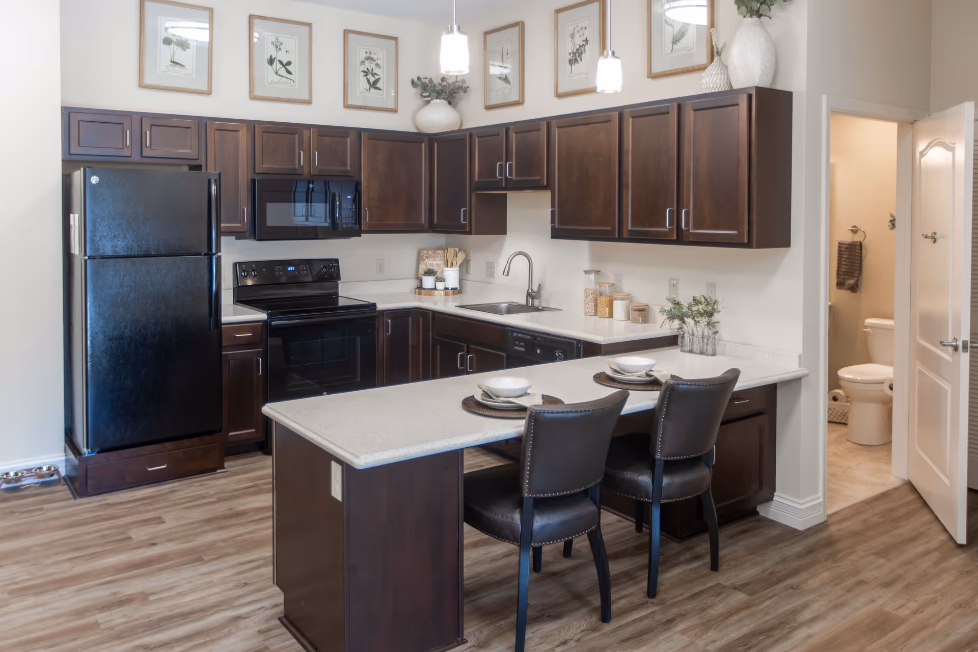 A modern kitchen with dark wood cabinets, black appliances including a refrigerator, oven, and microwave. There is a light-colored countertop island with two place settings and two dark chairs. The kitchen has wood flooring and framed botanical prints on the wall above the cabinets. A doorway to a bathroom with a toilet and towel rack is visible on the right.