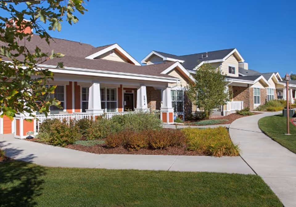 Single-story senior living building with covered porches, landscaped gardens and a curved walkway under a clear blue sky.