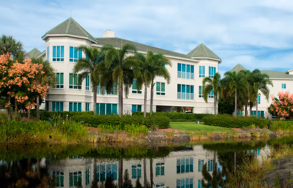 Three-story white building with turrets, palm trees, and a pond reflecting the facade.