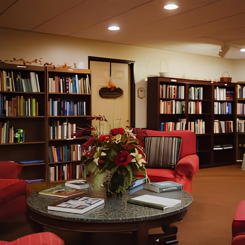 A cozy library or reading room with bookshelves filled with books along the walls. In the center, there is a round table with a floral arrangement and several books on it. Surrounding the table are red upholstered armchairs with striped cushions. The room has warm lighting and a door in the background.
