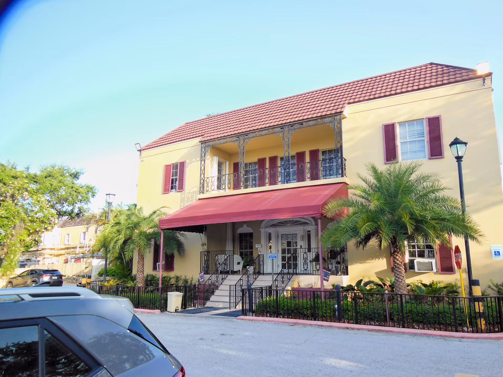 Two story yellow senior living building with red awning, balcony, palm trees, and parked cars