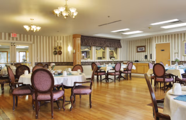 A dining room in a senior living facility with round tables covered in white tablecloths, set with cups and plates. The room has wooden flooring, striped wallpaper, and multiple chairs with purple upholstery. There are chandeliers on the ceiling and a counter area with cabinets along one wall.