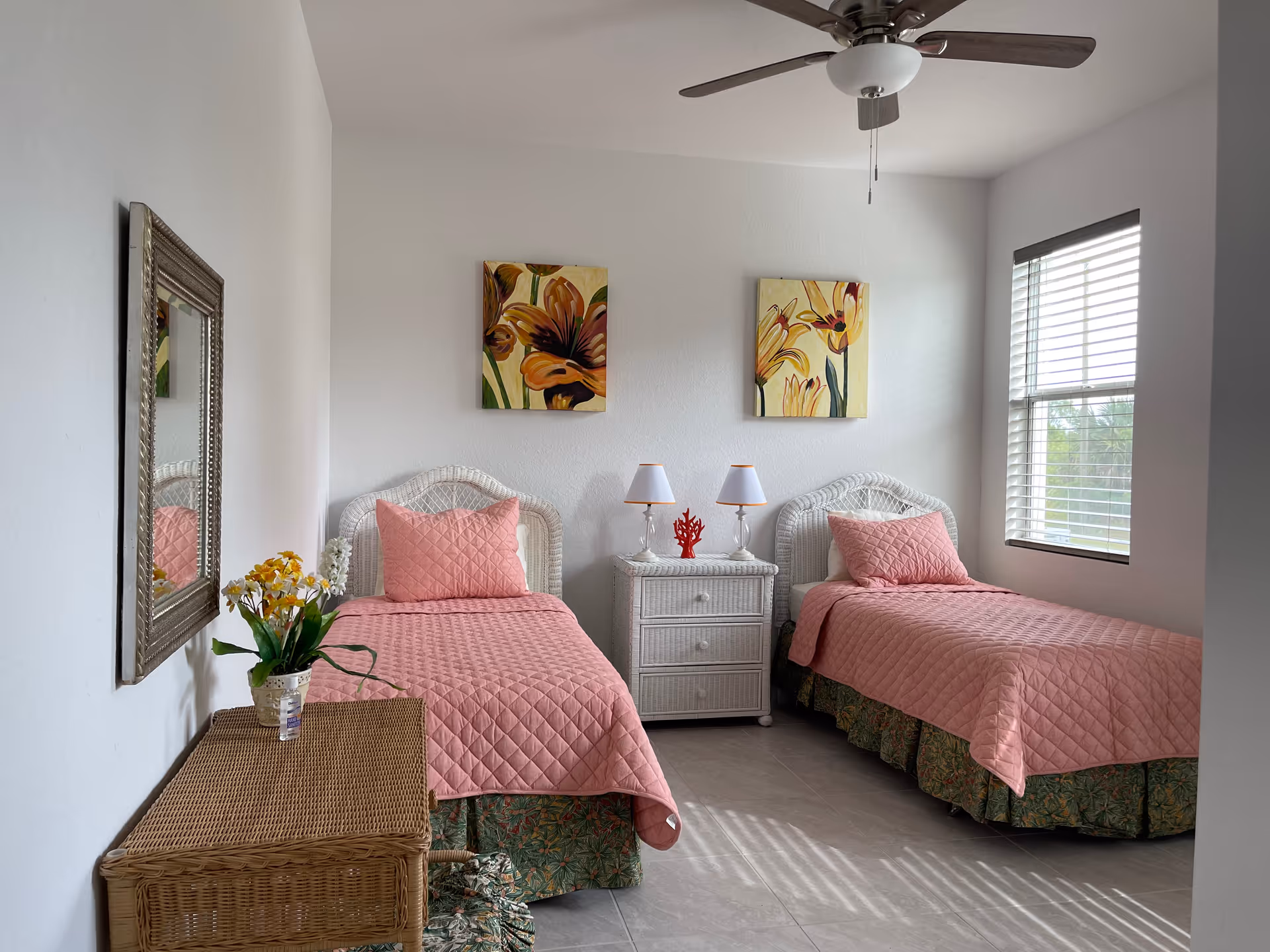 Two twin beds with pink quilts, white wicker headboards, a shared nightstand, and sunlight.