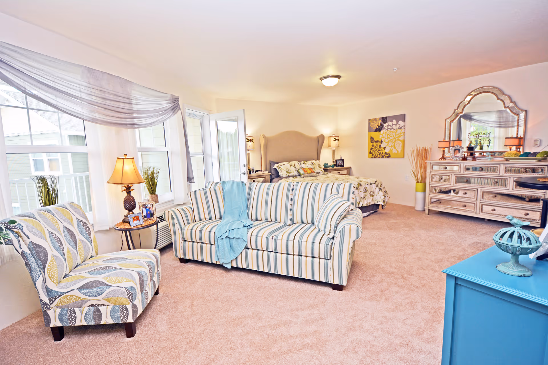A bright and cozy room featuring a striped sofa with a blue throw, a patterned armchair, a bed with a beige headboard and floral bedding, a large decorative mirror above a dresser, and a blue cabinet. The room has beige carpet, large windows with sheer curtains, and soft lighting from lamps.
