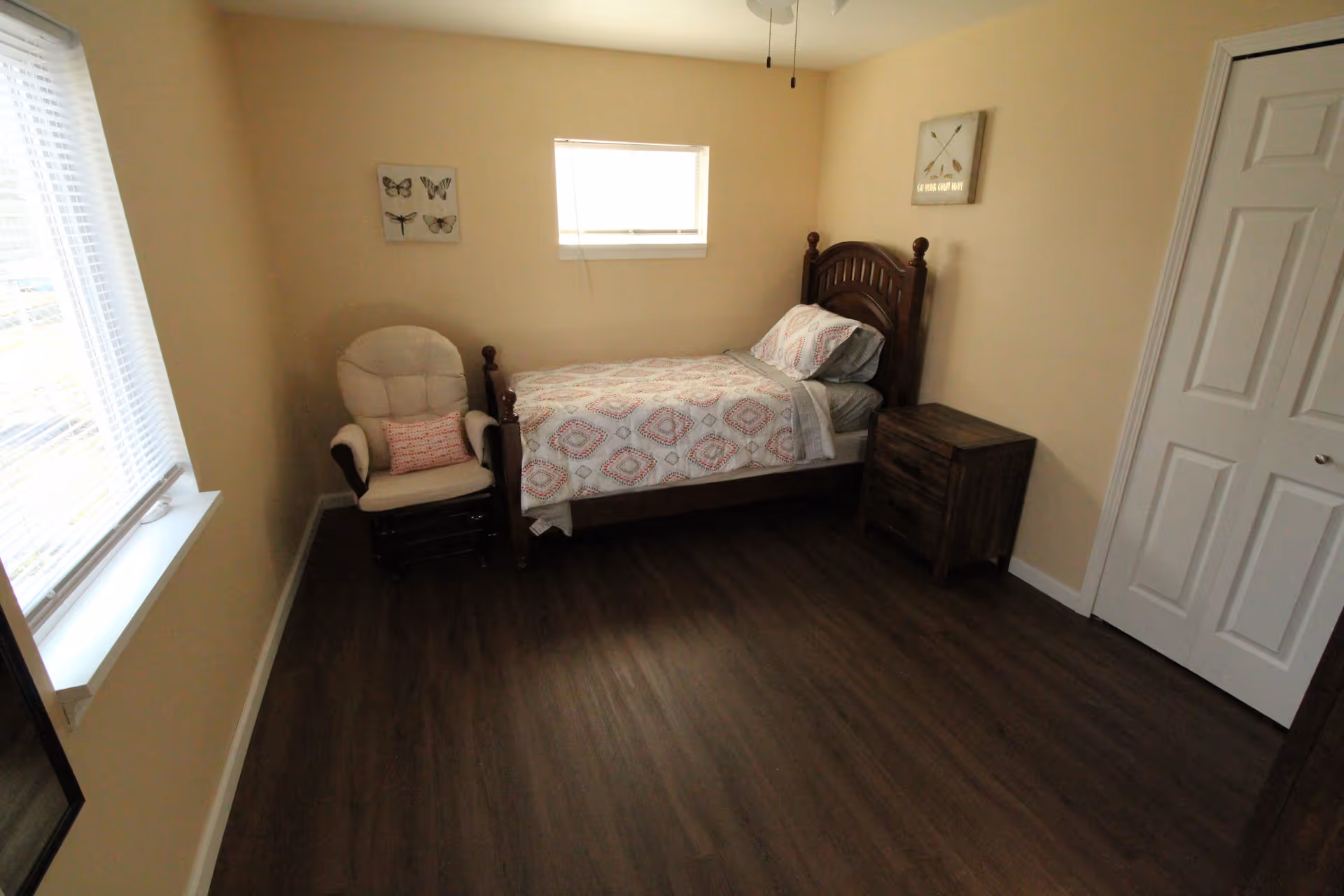 Small, tidy bedroom with a single wooden bed, nightstand, upholstered rocker, and windows.