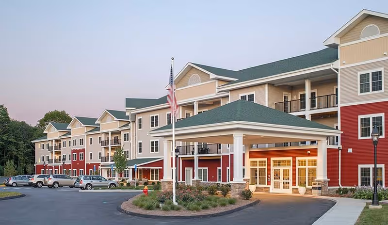 Exterior view of a multi-story senior living facility building with beige and red siding, green roofs, and a covered entrance. Several cars are parked in front, and an American flag is displayed on a flagpole near the entrance. Trees and landscaping surround the building.