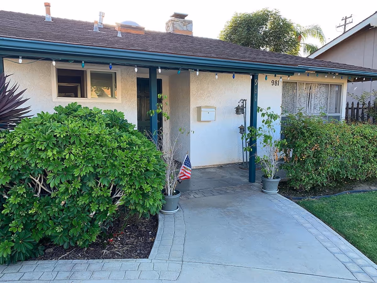 Front entrance of a single-story house with a covered porch, green shrubs, potted plants, and a small American flag by the walkway.