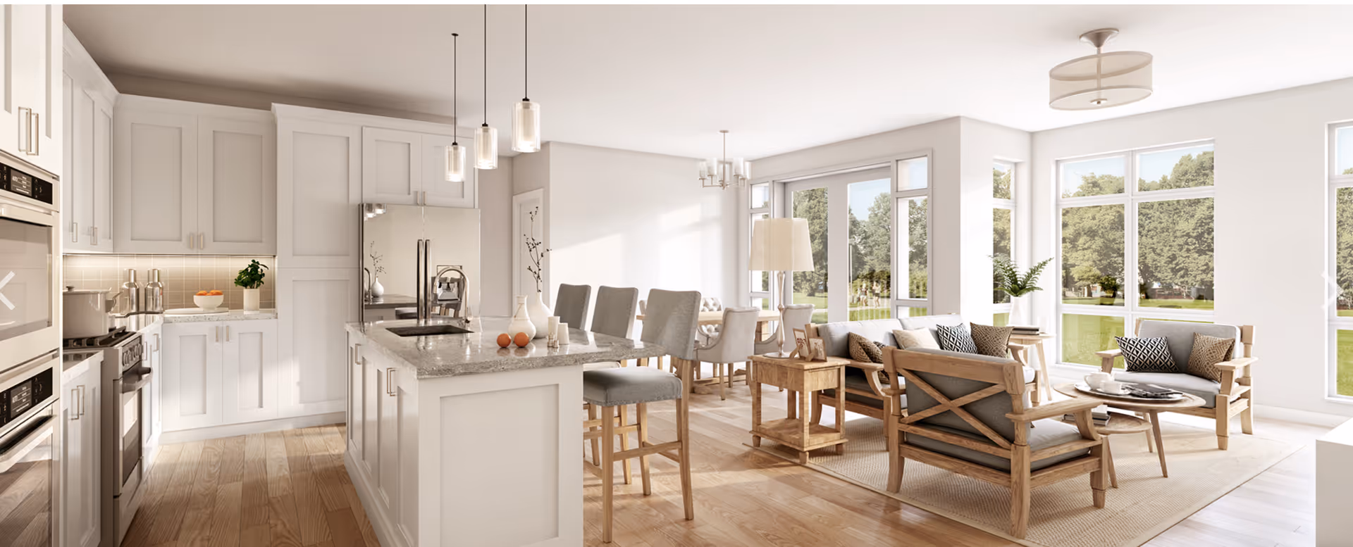Bright open-concept kitchen and living area with white cabinetry, a marble island with stools, a dining table, and wood-framed lounge chairs by large windows overlooking a green lawn.