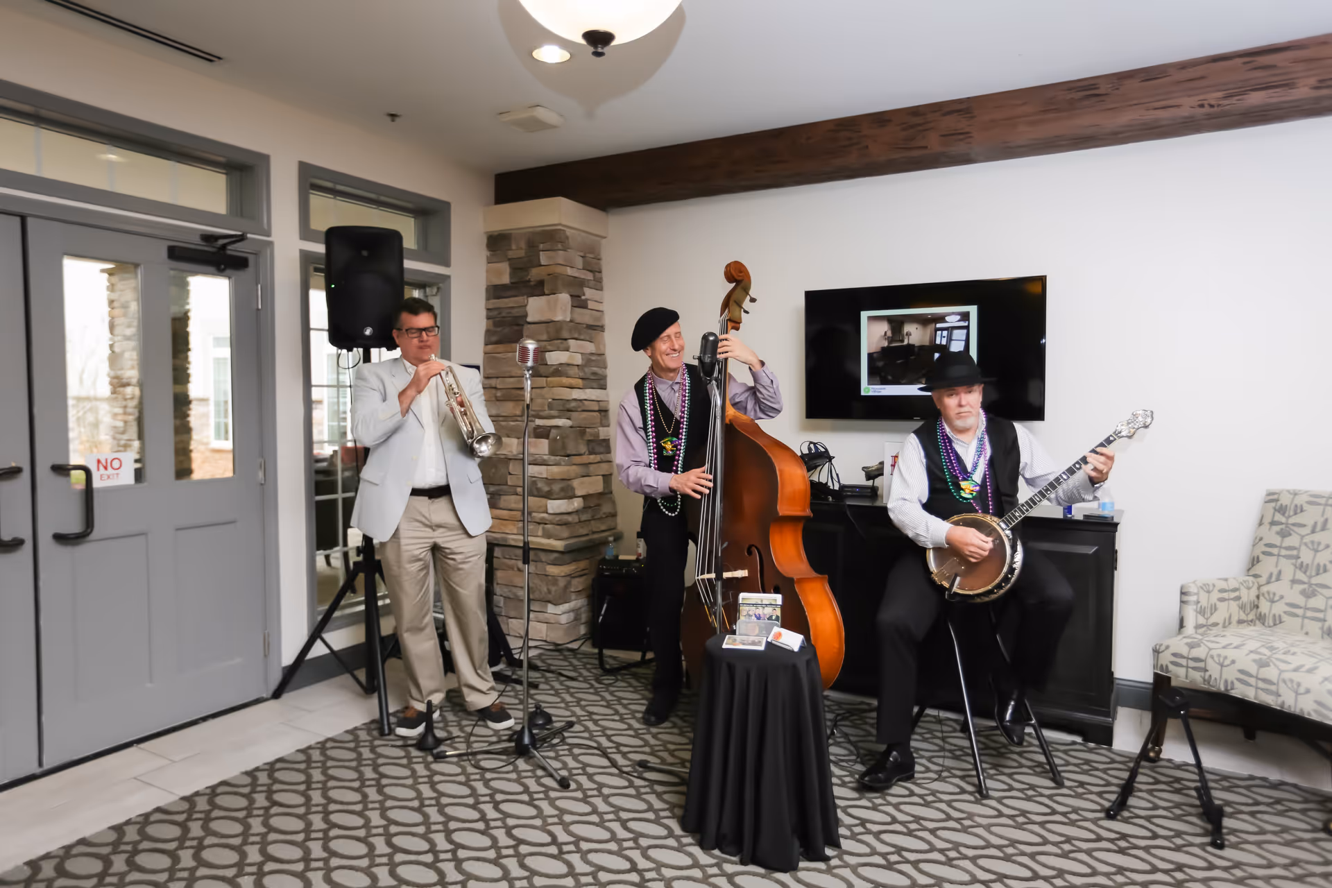 Three musicians performing indoors at The Landings of Canton Hills. One man is playing a trumpet, another is playing a double bass, and the third is playing a banjo. They are standing or sitting on a patterned carpeted floor near a stone pillar and a TV mounted on the wall. The room has a modern interior with a door labeled 'NO EXIT' and a patterned armchair to the right.