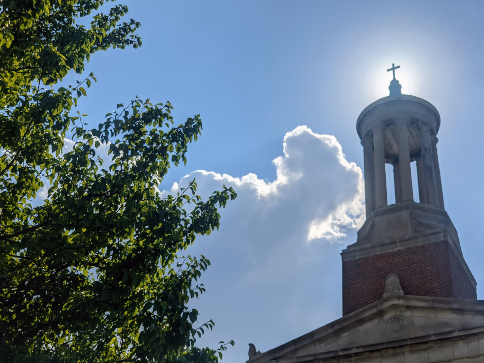 Bright sky, tree branches frame a tall church tower with a cross on top.