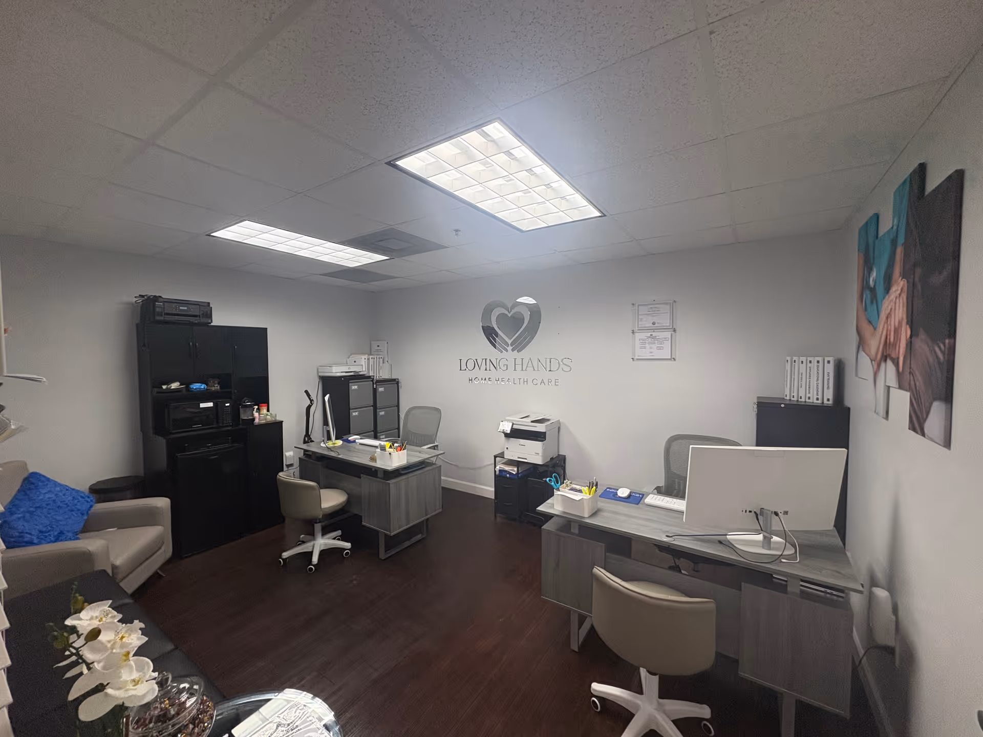 Office reception area with desks, chairs, printers, and Loving Hands Home Health Care logo on wall.
