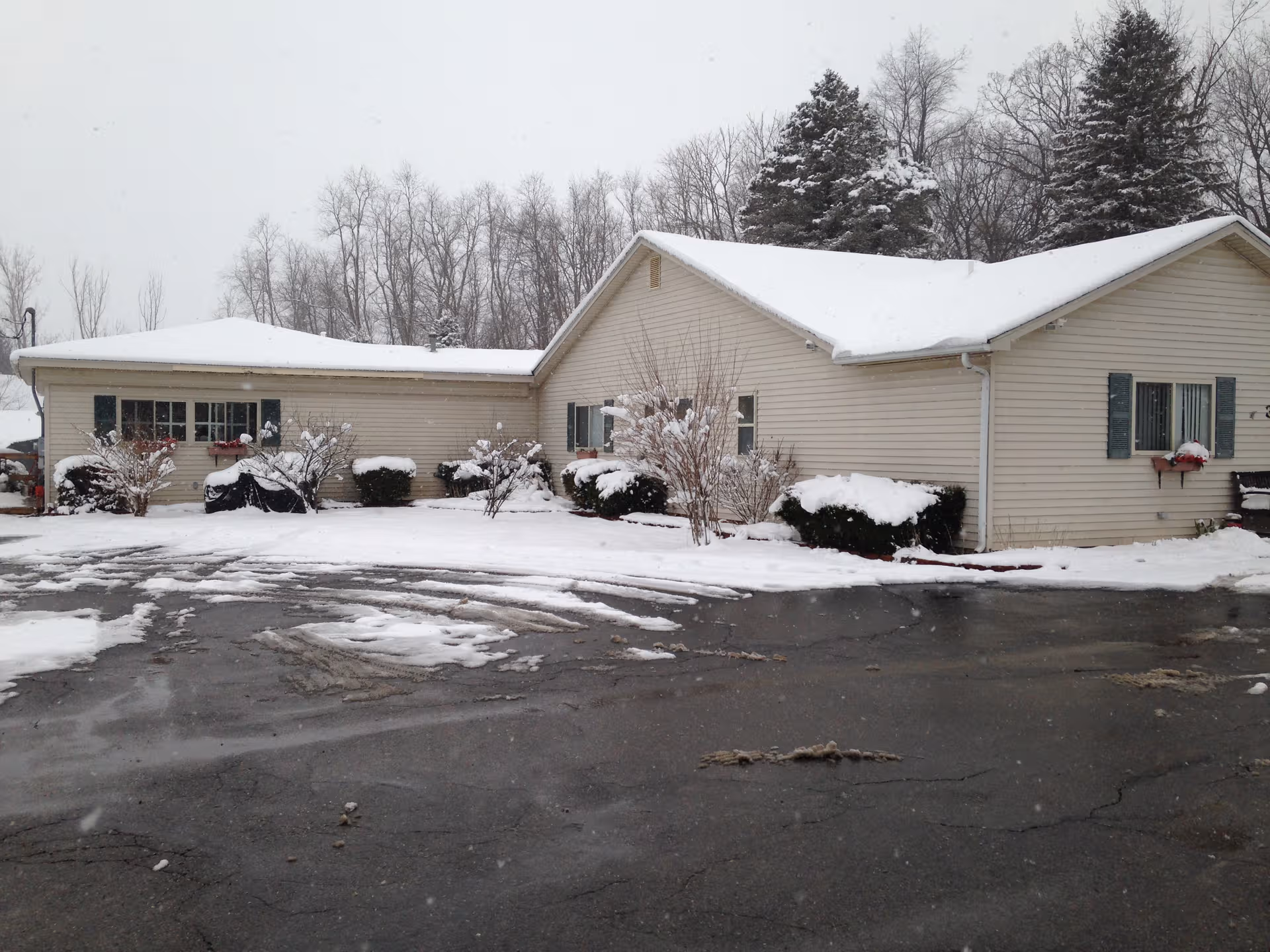 Snowy, single-story beige building with gabled roof, shrubs, and a wet parking lot in winter.