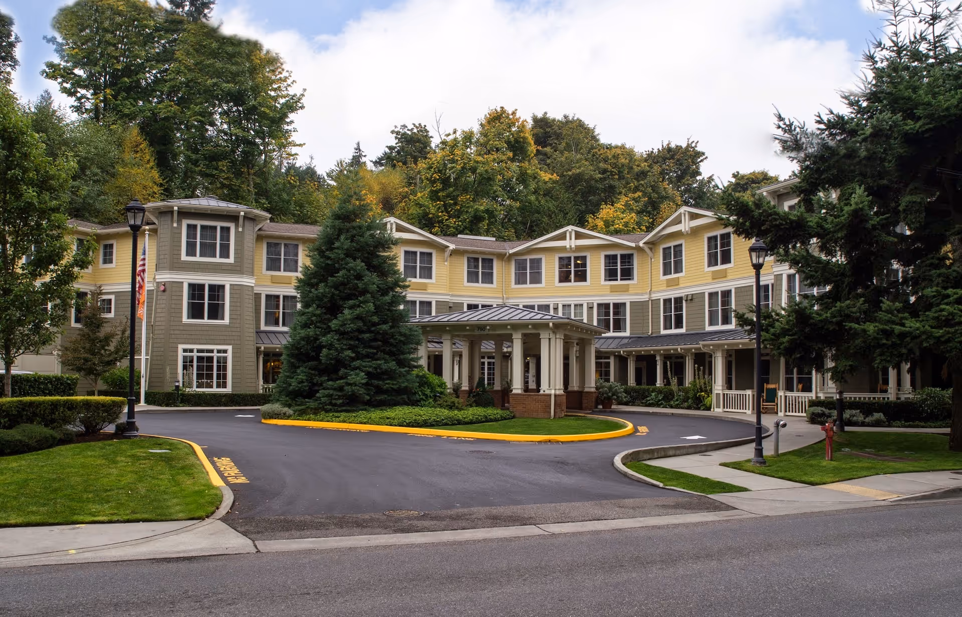 Exterior view of a three-story senior living facility building with a covered entrance driveway, surrounded by well-maintained landscaping including trees, bushes, and grass. The building features a combination of yellow and gray siding with multiple windows and a backdrop of tall trees under a partly cloudy sky.