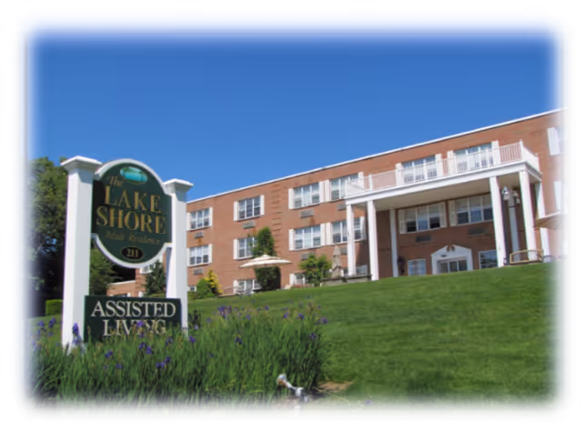 Exterior view of a three-story brick assisted living facility named The Lake Shore Adult Residence, with a green lawn and purple flowers in the foreground under a clear blue sky.