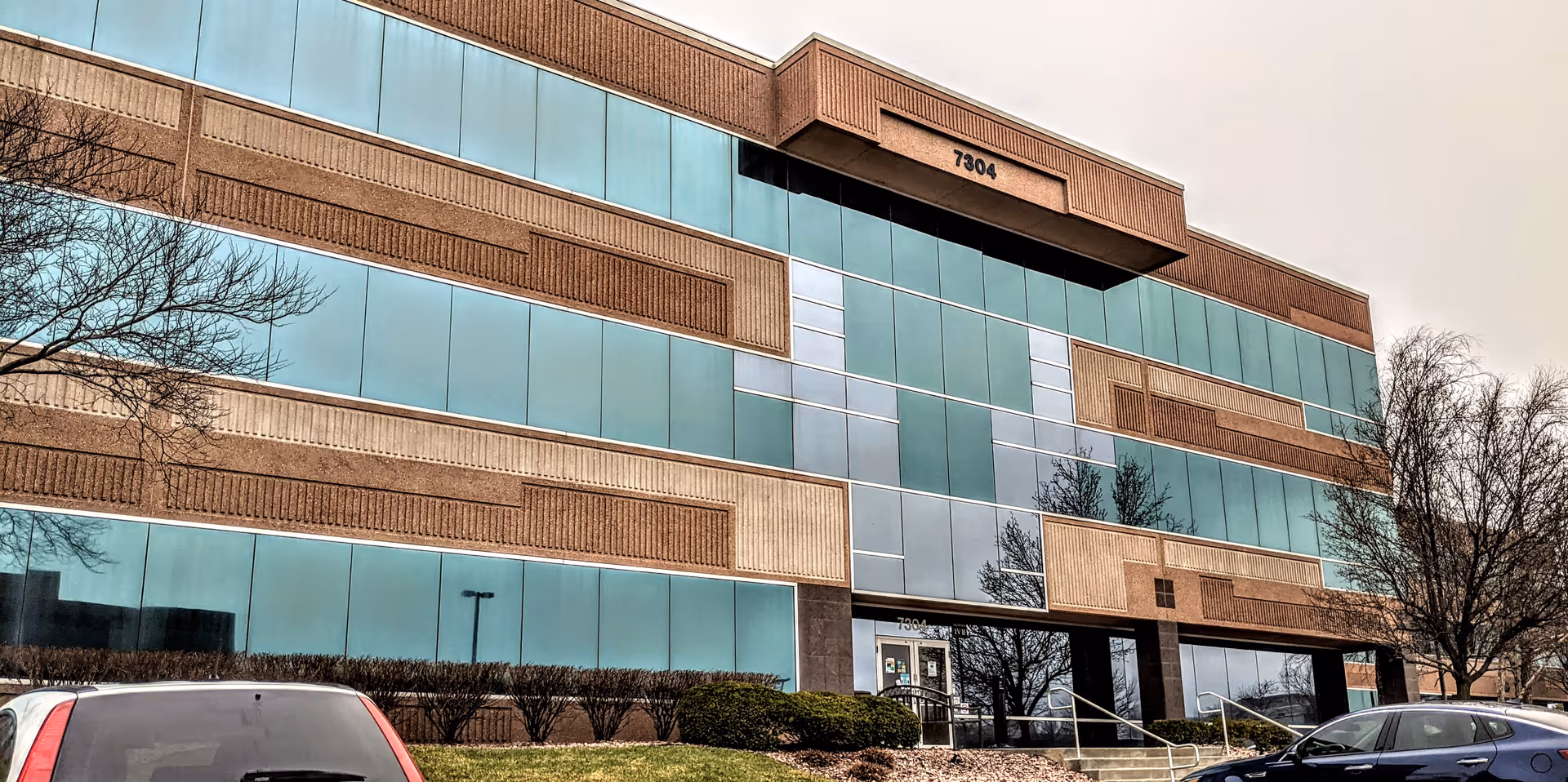 Front exterior view of brick and glass senior living facility with entrance and parked cars.