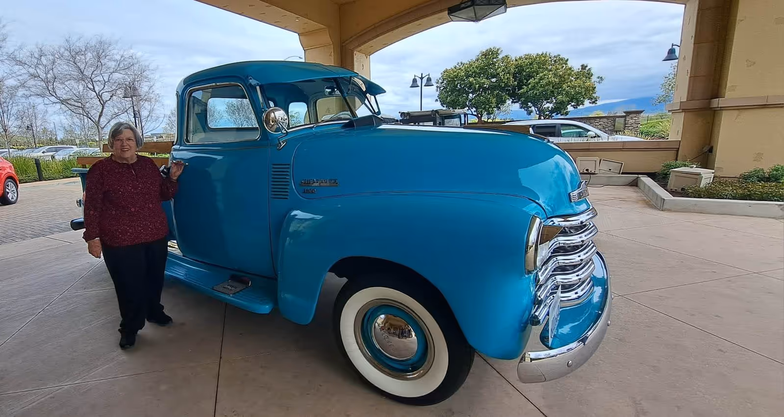 An elderly woman stands beside a vintage blue truck under a covered facility entrance.