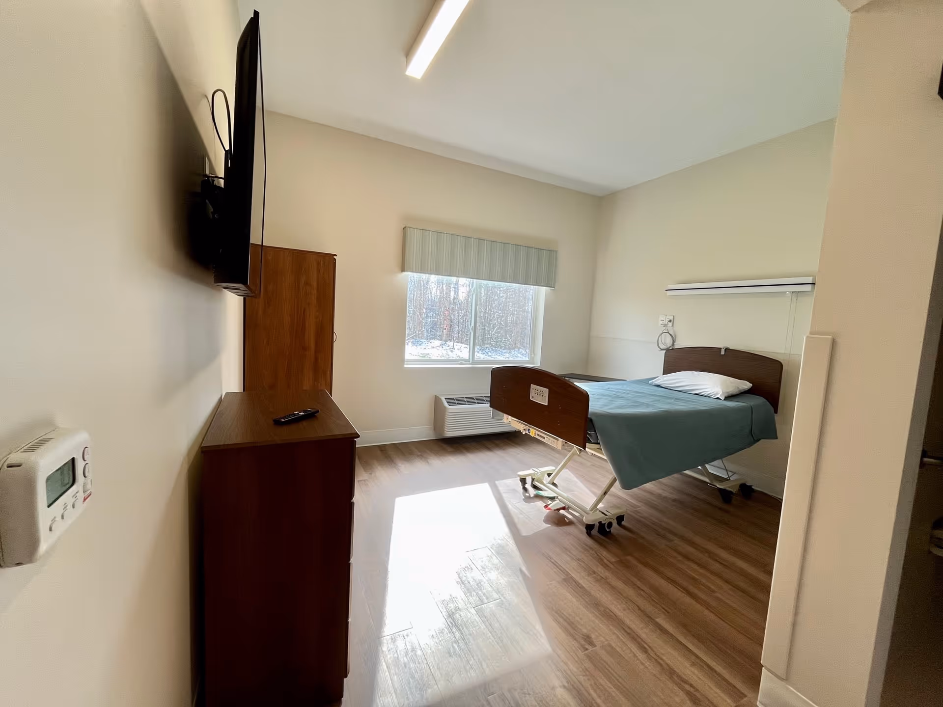 A senior living patient room with a single bed, dresser, wall-mounted TV, and window.