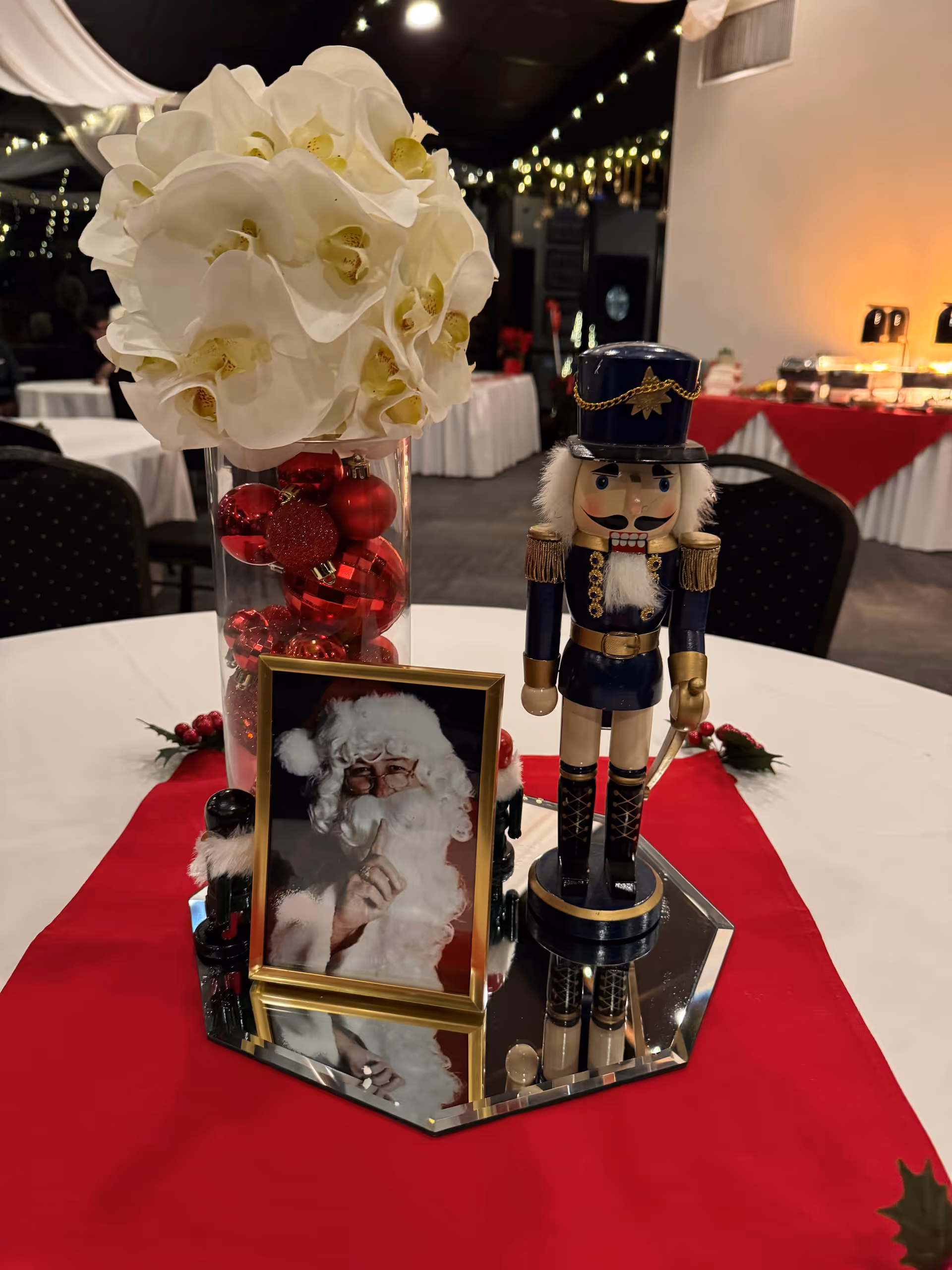 Holiday table centerpiece with white flowers, a nutcracker, and Santa photo on a red cloth.