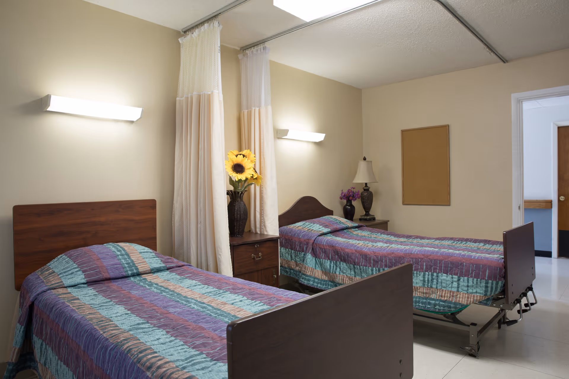 A room with two single beds separated by a wooden nightstand with a vase of sunflowers. Each bed has a colorful striped bedspread. There are privacy curtains hanging from the ceiling between the beds. The room has beige walls, a corkboard on one wall, a table lamp, and a door leading to another area.
