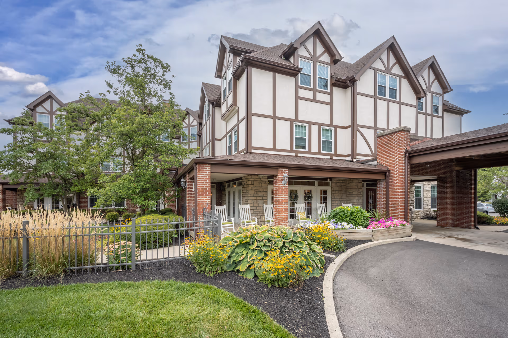 Entrance of a multi-story Tudor-style senior living building with a covered drive, rocking chairs on the porch, and landscaped flower beds.