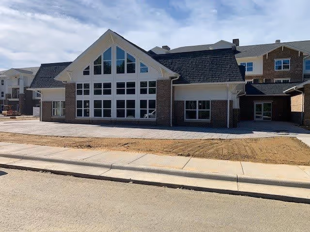 Exterior view of a large brick building with multiple windows and a steeply pitched roof under a partly cloudy sky. The building appears to be a senior living facility with a paved sidewalk and a dirt area in front.