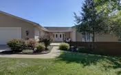 Single-story assisted living facility front with a lawn, walkway, shrubs, double front doors, and an attached garage under a clear sky.
