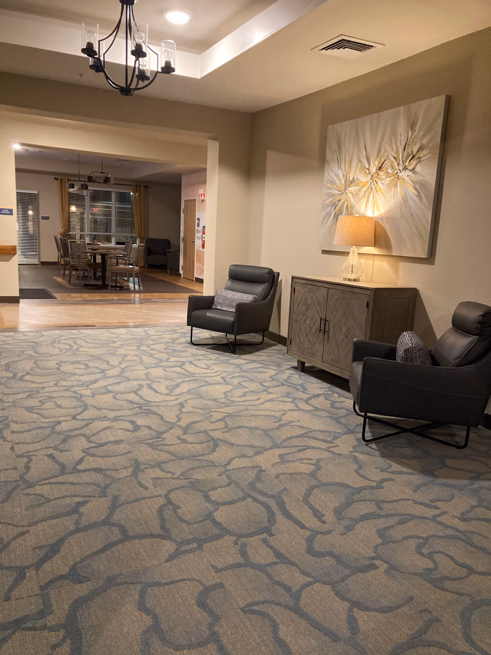 Warm lobby seating area with two dark armchairs, cabinet, artwork, and a distant dining area.