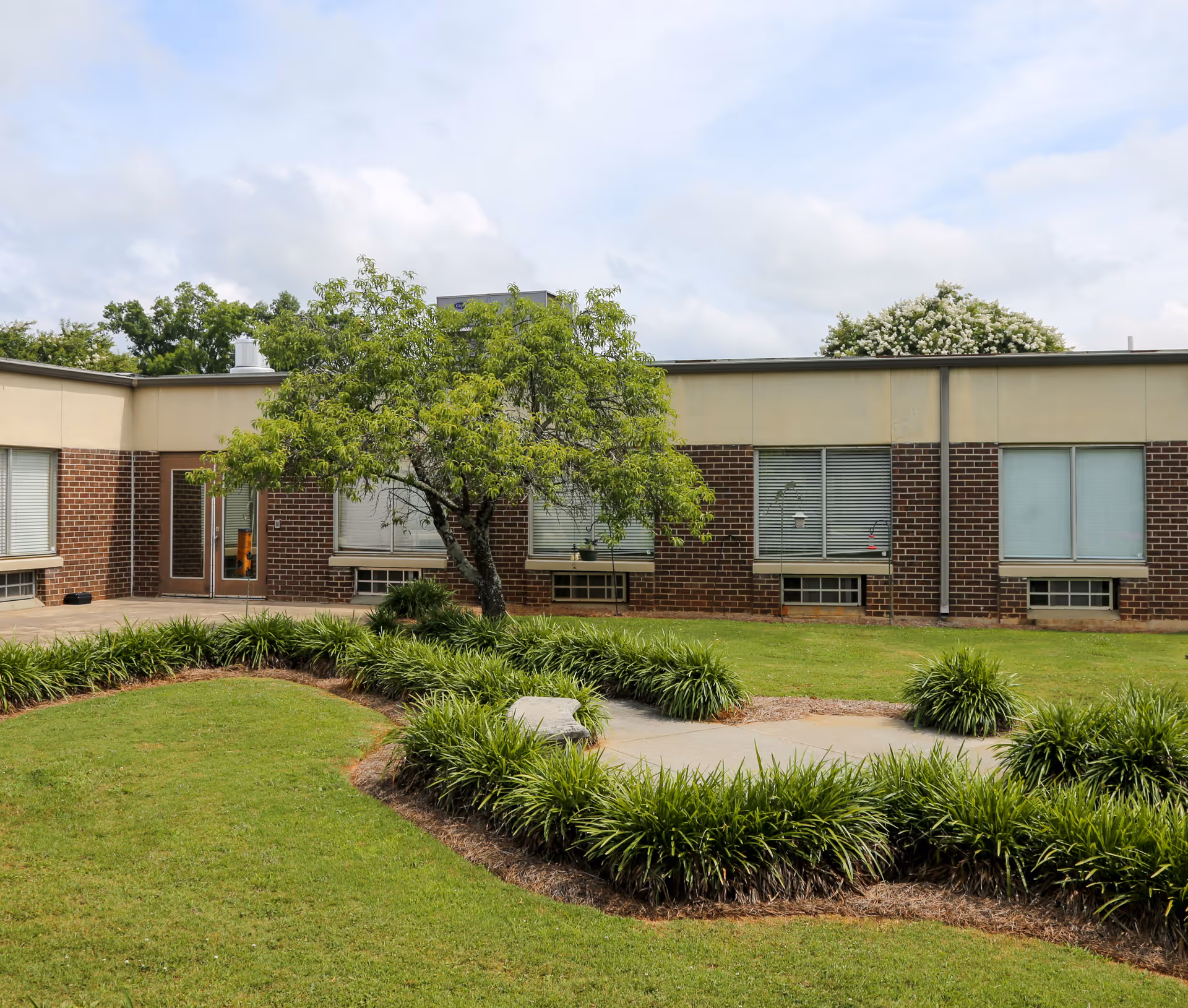 Exterior view of a single-story brick facility with landscaped circular walkway and trees.
