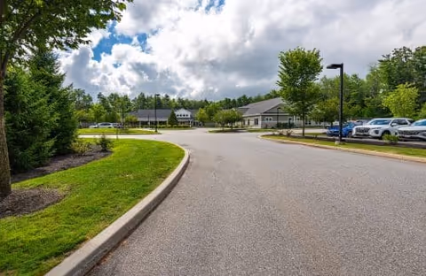 Wide paved driveway leading to a single-story building surrounded by green trees and grass under a partly cloudy sky. Several parked cars are visible on the right side of the driveway.