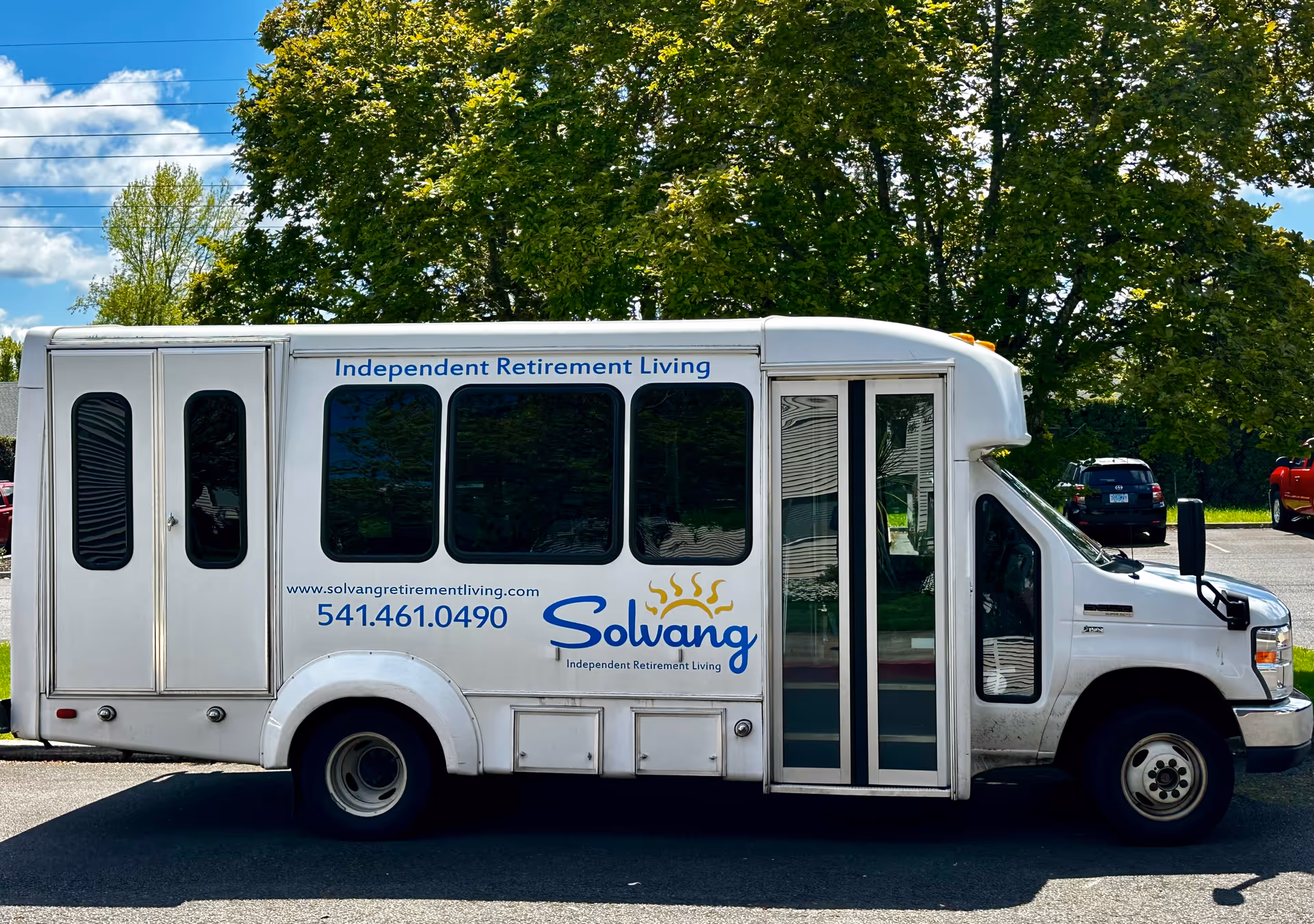 White shuttle van branded 'Solvang Independent Retirement Living' parked in a lot with trees in the background.