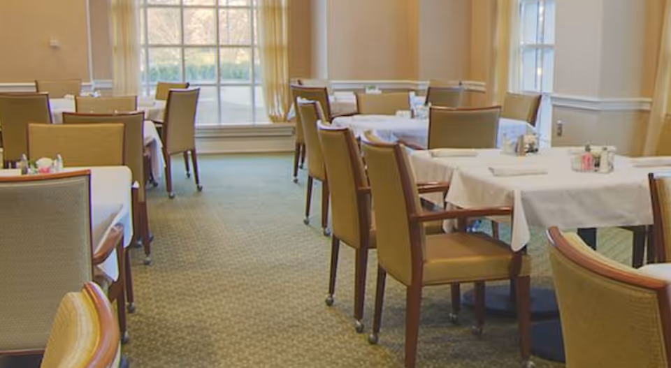 A dining room with multiple tables covered in white tablecloths and surrounded by wooden chairs with cushioned seats. The room has large windows with light curtains allowing natural light to enter.