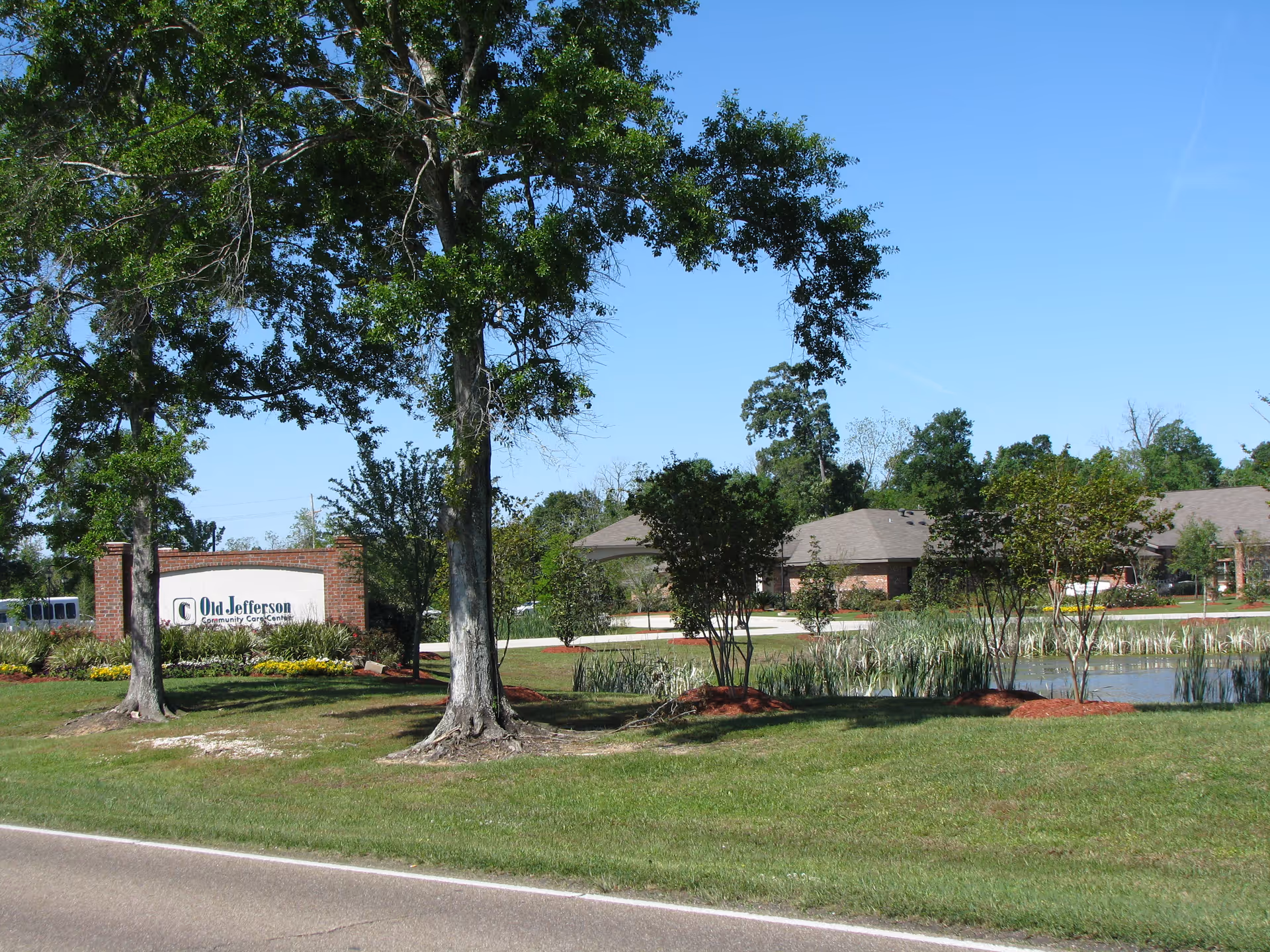 Front exterior of Old Jefferson Community Care Center showing the entrance sign, trees, pond, and single-story brick buildings under a clear blue sky.